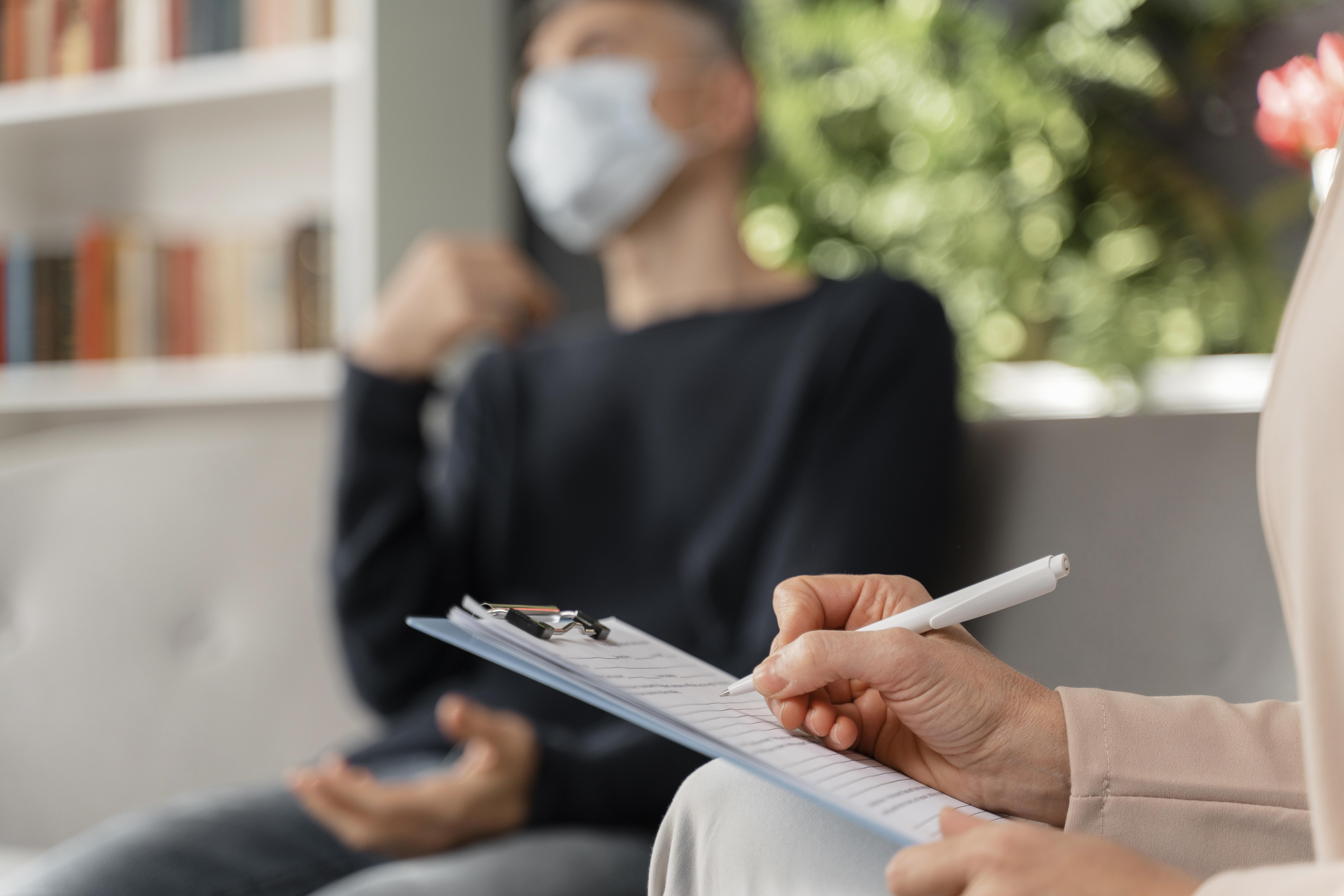 woman-therapist-taking-notes-of-man-in-therapy-office-with-mask.jpg